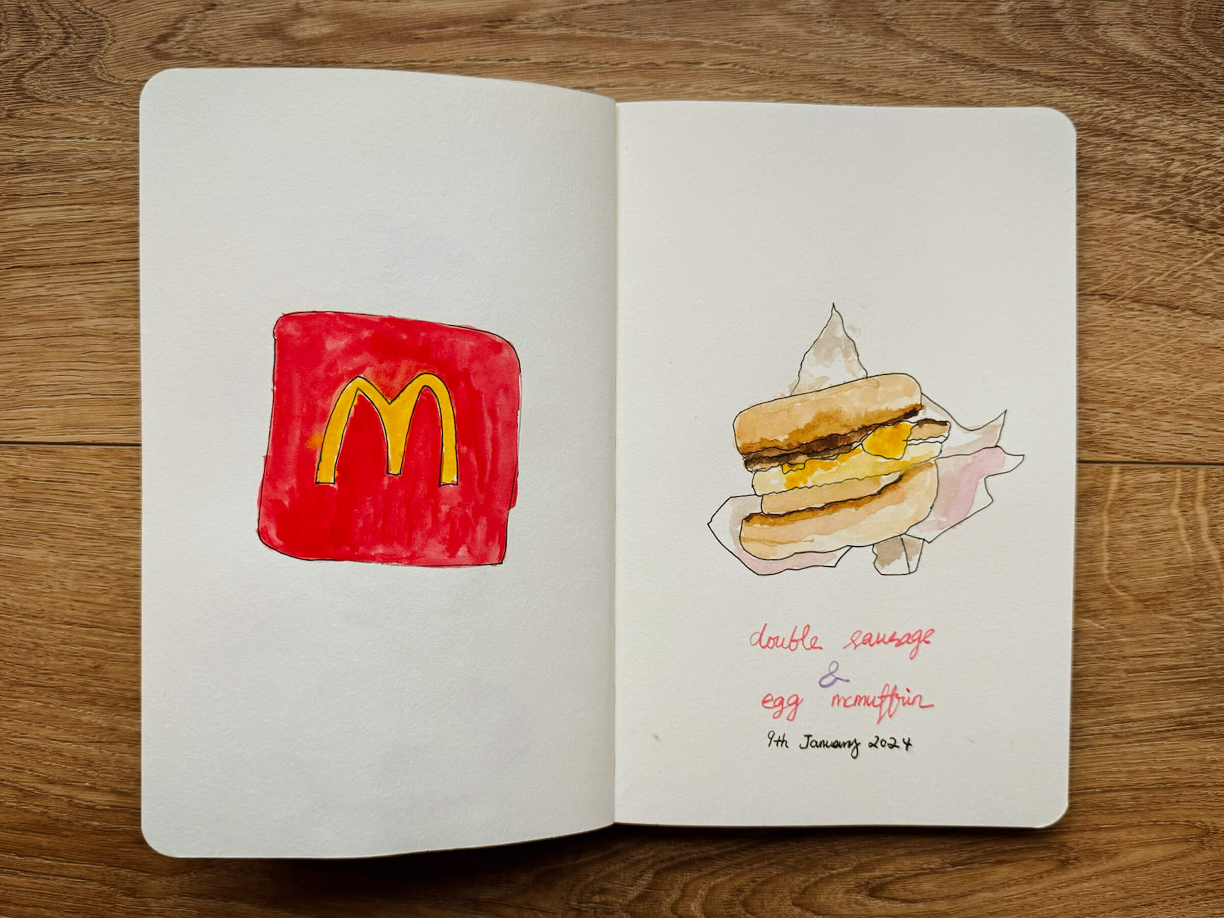 photo of my sketchbook with a spread containing a painting of macdonald's logo with a double sausage & egg mcmuffin. drawn on 9th jan 2024.